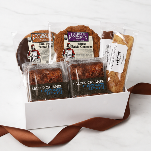 A gift box of cookies and brownies from Cougar Mountain Bakes, including Dark-Chocolate Peanut Butter Cookies, Oatmeal Raisin Cinnamon Cookies, Salted Caramel Chocolate Brownies, and a Chocolate Chip Cookie, arranged in a white box with a brown ribbon on a white marble background.