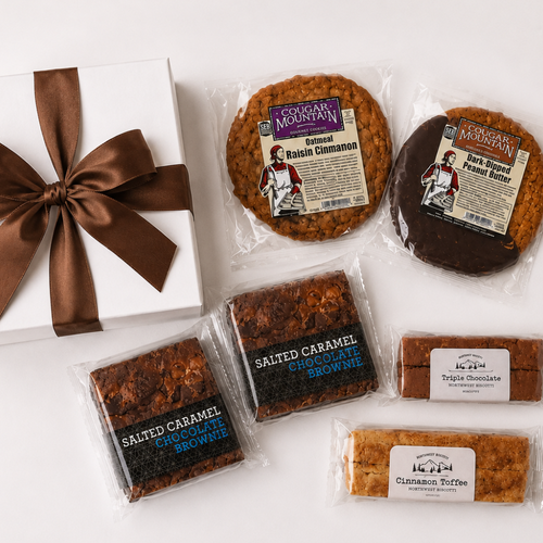 A gift assortment of cookies and brownies from Cookie Monster's Mountain, including packaged salted caramel chocolate brownie, salted caramel chocolate cookie, salted cinnamon cinnamon, and triple chocolate cookie, arranged on a white surface next to a white gift box with a brown ribbon.
