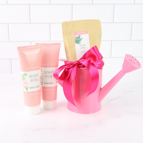 Pink watering can with a bow, body wash, and lotion on a white tiled background
