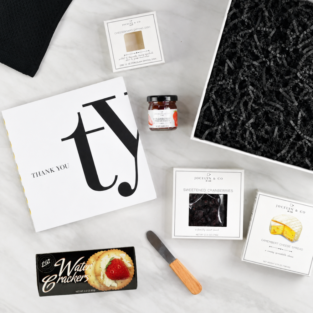 A curated gift box set from Jessica & Co. arranged on a white marble surface, featuring a white 'Thank You' card, a jar of preserves, water crackers, cranberry sauce, cheese spread, a small knife, and a black textured insert inside a gift box.