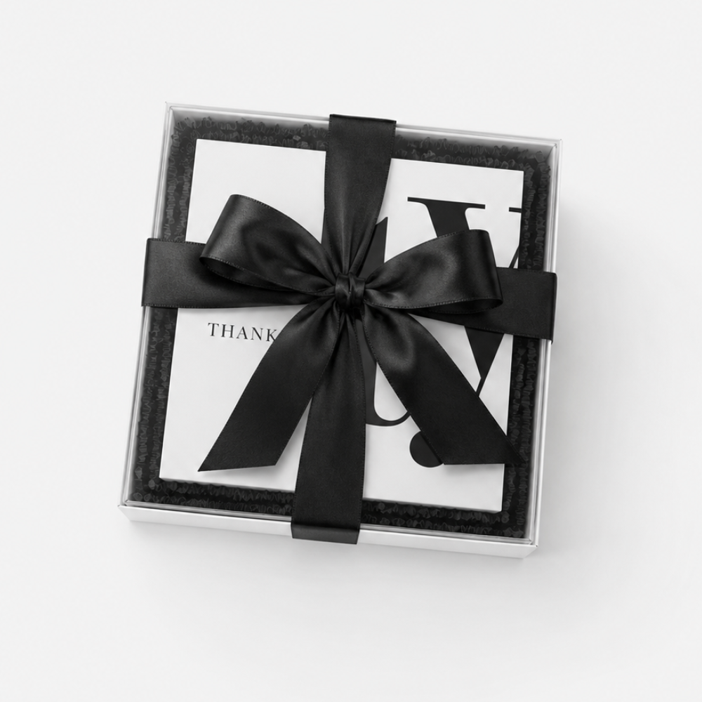 A square gift box with a clear lid, lined with black velvet, tied with a wide black satin ribbon bow. A white card with partial black lettering is visible inside.