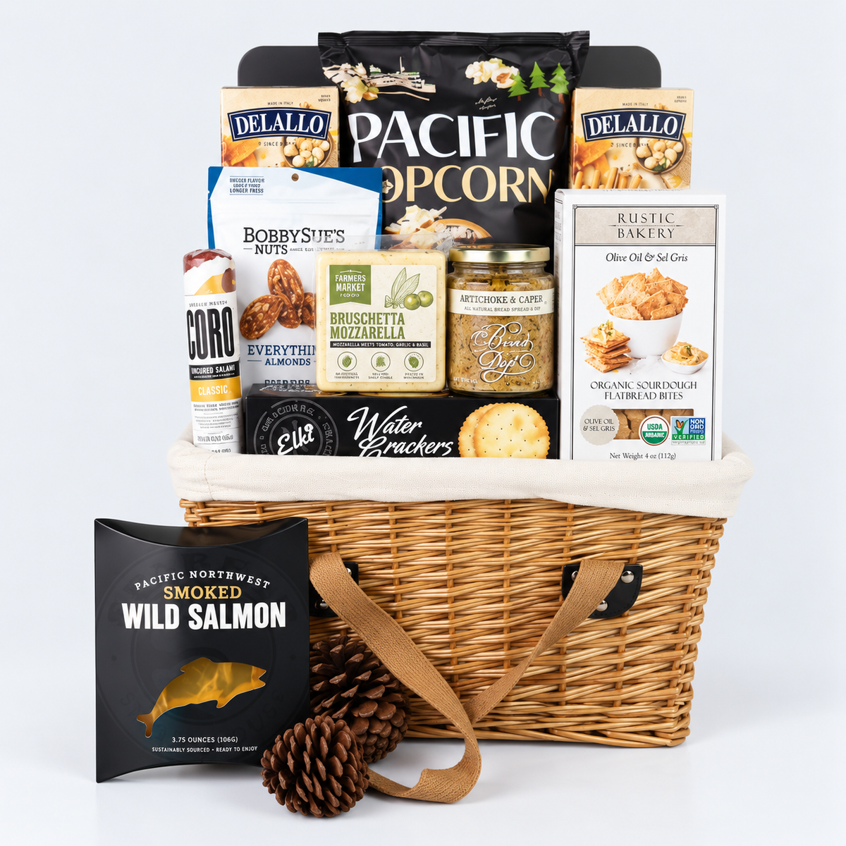 A wicker gift basket filled with assorted snack and food items, including Delallo popcorn, Bobbie's Bites, Rustic Bakery cookies, and a Pacific Northwest smoked wild salmon packet, arranged on a white background with two pinecones in front.